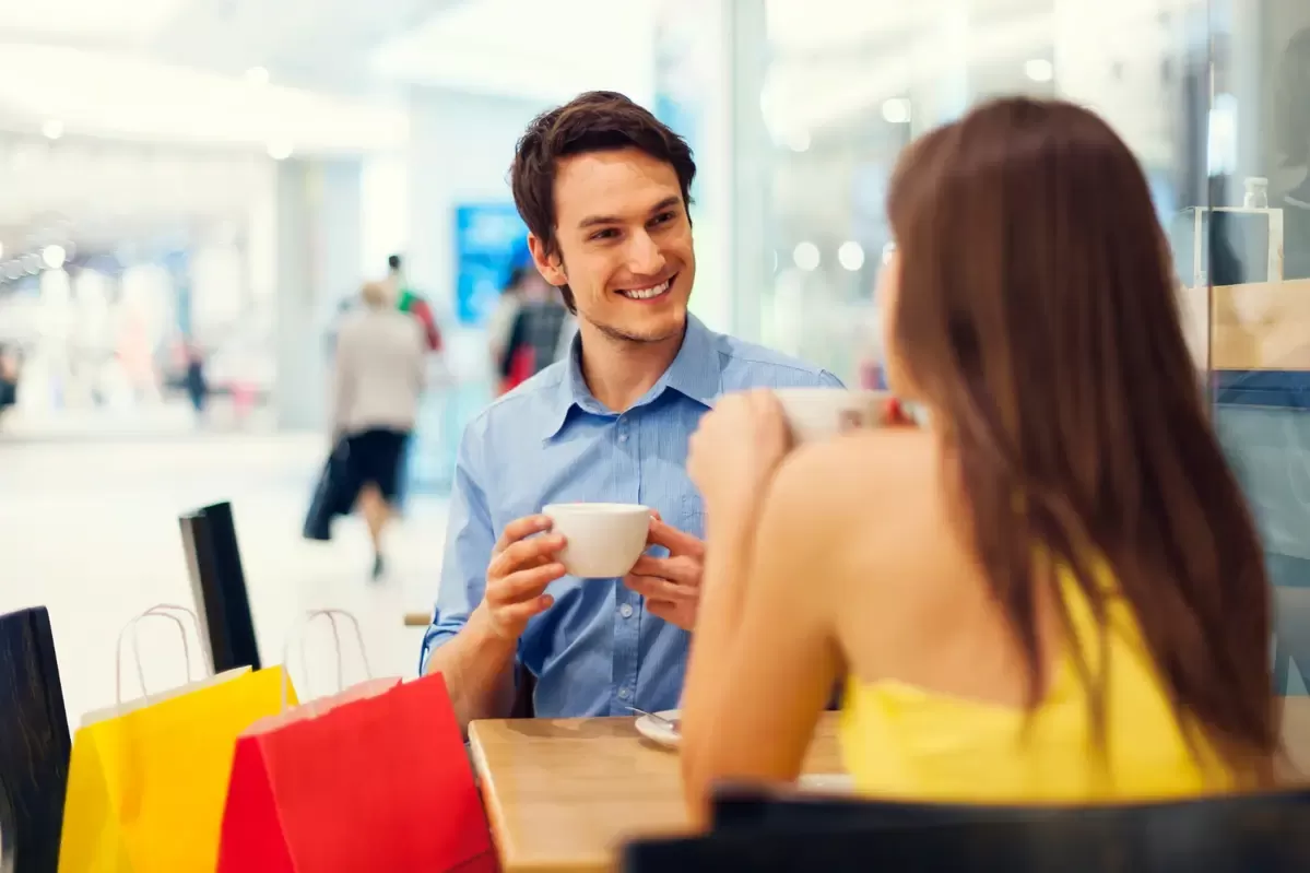 Homem de camisa azul e mulher de vestido amarelo conversam sorrindo enquanto tomam café em uma cafeteria dentro de um shopping, representando franquias baratas para shopping. Sacolas de compras coloridas estão em uma cadeira ao lado.