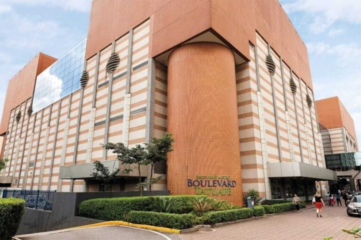 fachada do Shopping Boulevard Tatuapé. O prédio é de estilo moderno, com um design que combina paredes de tijolos e grandes superfícies de vidro. Na parte central, há um pilar circular em tom laranja, que se destaca no conjunto. O nome do shopping está visível na base da fachada, em letras verdes: "Boulevard Tatuapé". O entorno do shopping é bem cuidado, com áreas ajardinadas e uma rua pavimentada.