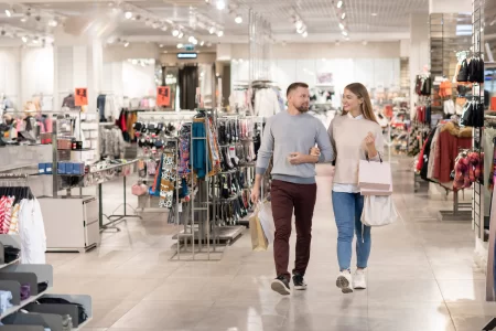 A imagem mostra um casal caminhando por um shopping, passando por várias prateleiras de roupas e acessórios, representando diferentes tipos de lojas em shopping.