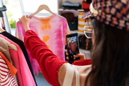 Mulher fotografando uma camiseta tie-dye em uma arara de roupas para divulgar em suas redes sociais. Uma das melhores ideias de marketing para atrair clientes.